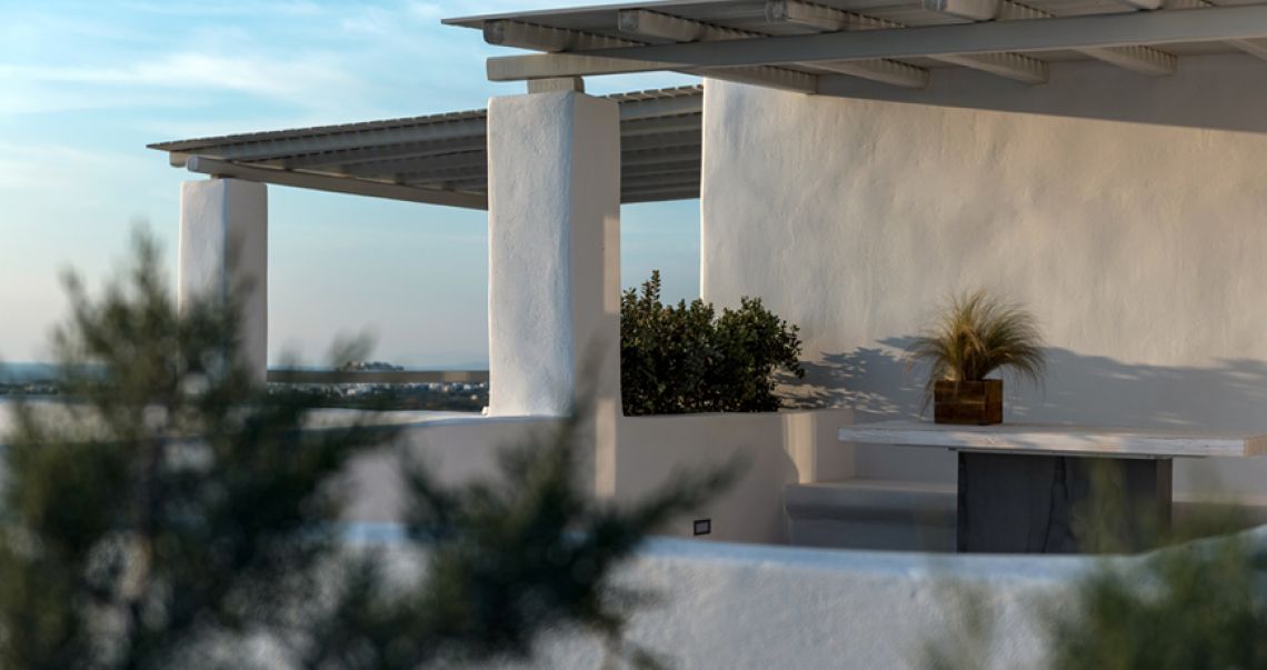 Golden hour view of a white Cycladic terrace with a wooden pergola, featuring a simple outdoor table decorated with a wooden planter against a soft sunset sky.