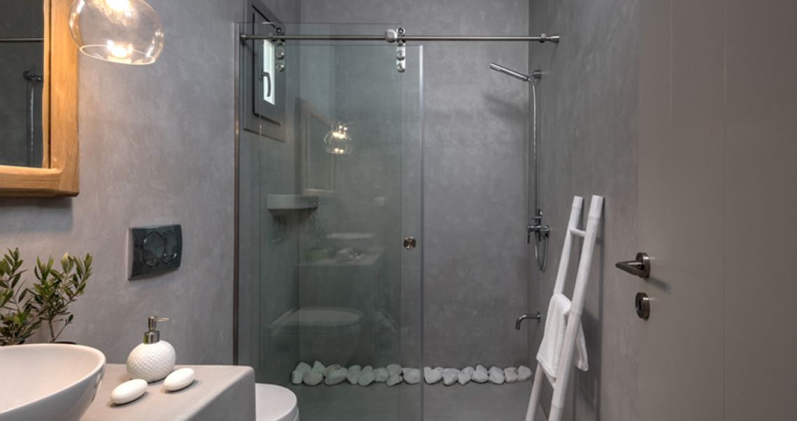 Modern grey microcement bathroom featuring a glass walk-in shower, a white vessel sink, and a decorative wooden ladder towel rack.