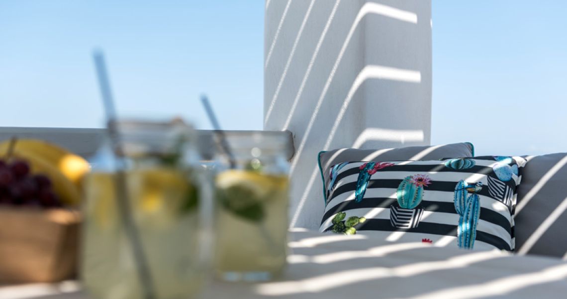 Sunny outdoor terrace detail featuring a cactus-print cushion and refreshing drinks in glass jars, cast with pergola shadows.
