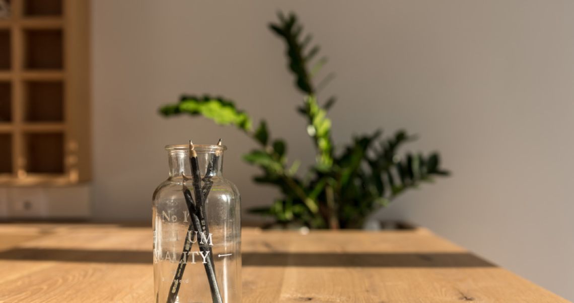 Close-up of a glass jar holding black pencils on a wooden desk, with a blurred green plant in the sunlit background.