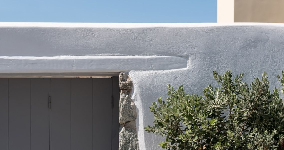 Close-up of traditional white stucco architecture featuring a grey wooden shutter, exposed stone detailing, and green foliage.