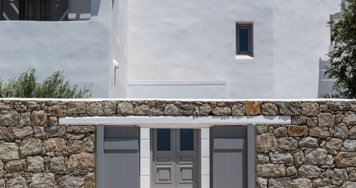 Exterior view of a traditional stone wall with grey double doors leading to a white Cycladic-style hotel building.