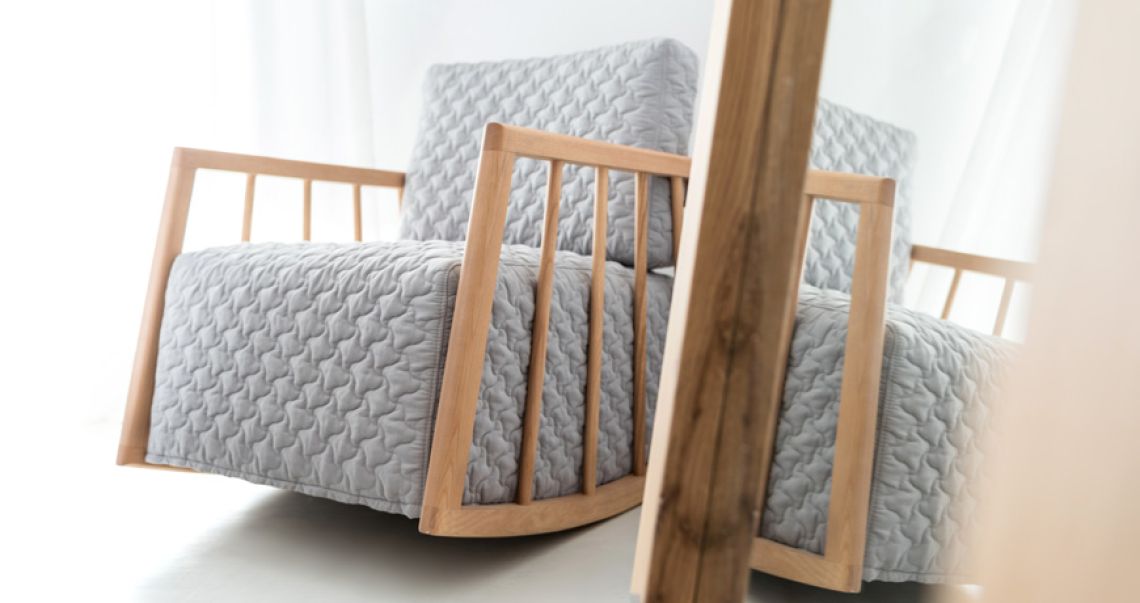 Modern grey quilted rocking chair with a light wooden frame, reflected in a mirror in a bright, airy room.