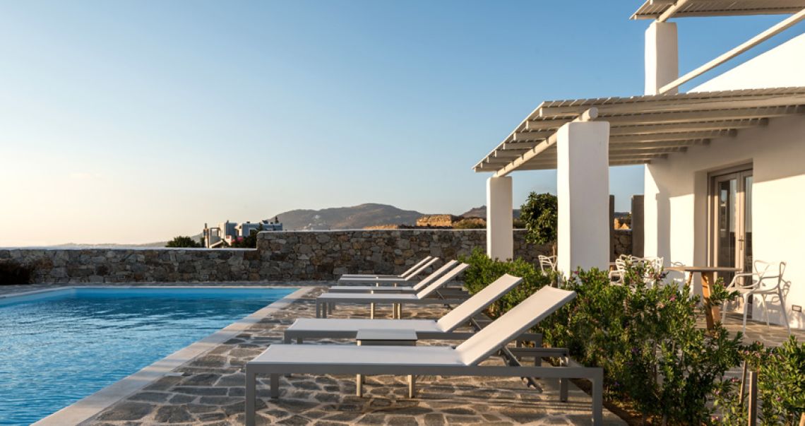 Outdoor swimming pool and stone patio with modern sun loungers, set against a white villa and scenic landscape during the day.