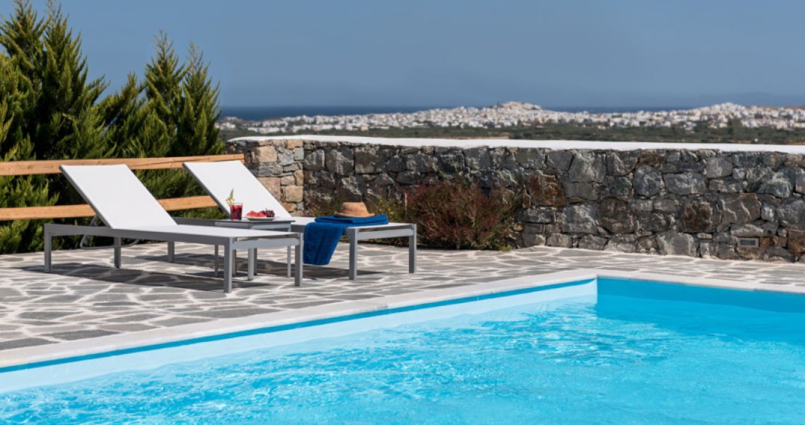 Two sun loungers with refreshments by the poolside, overlooking a stone wall and a panoramic view of the distant town and sea.