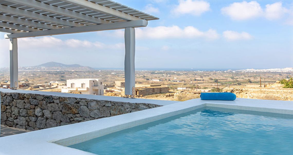 Eye-level view from a private plunge pool, looking out over a traditional stone wall towards a vast panoramic view of the island's countryside and sea.