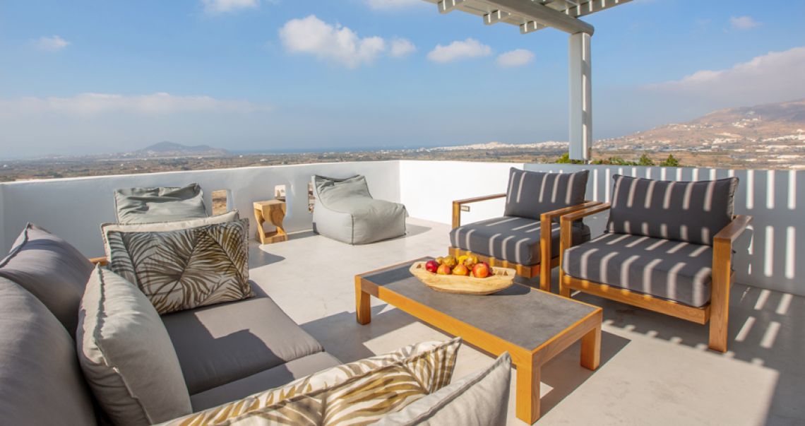 Sunlit outdoor lounge area under a pergola with comfortable grey seating and wide views of the countryside and distant mountains.