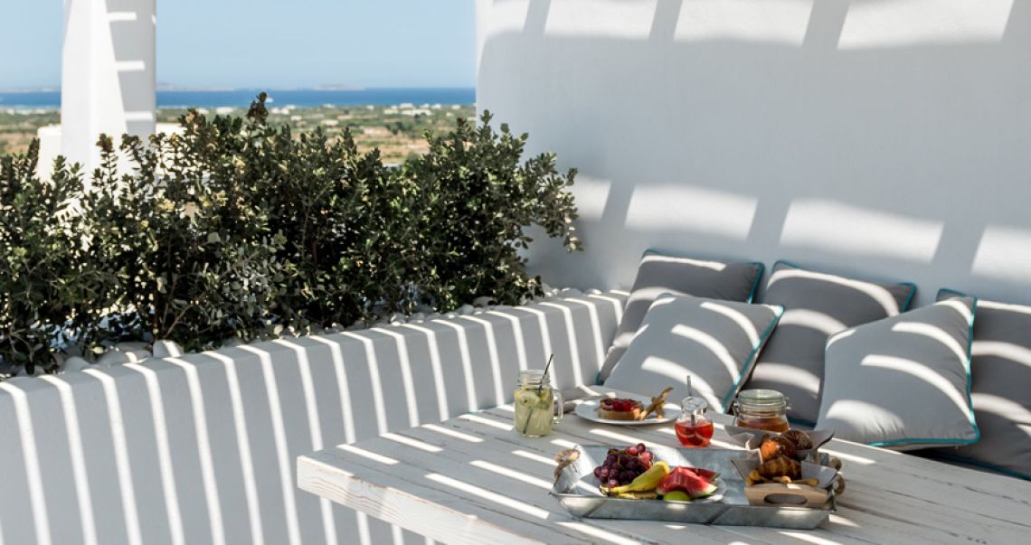 Sunlit outdoor breakfast setting on a white terrace, featuring a tray of fresh fruit and pastries on a wooden table, surrounded by grey cushioned seating and pergola shadows.