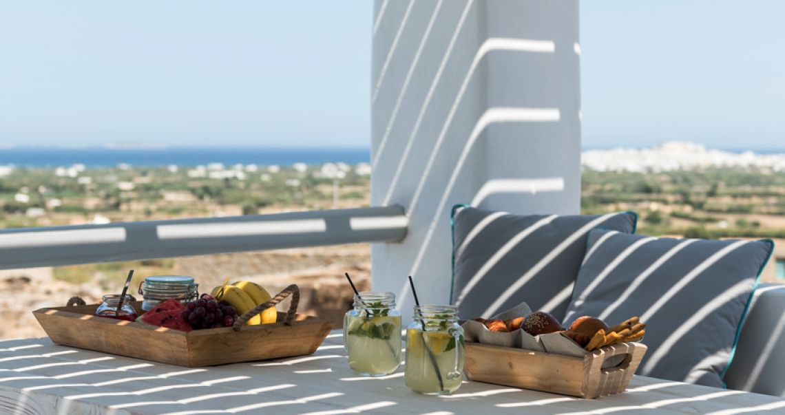 Sun-drenched balcony breakfast featuring fresh fruit, pastries, and lemonade, set against a backdrop of the distant sea.