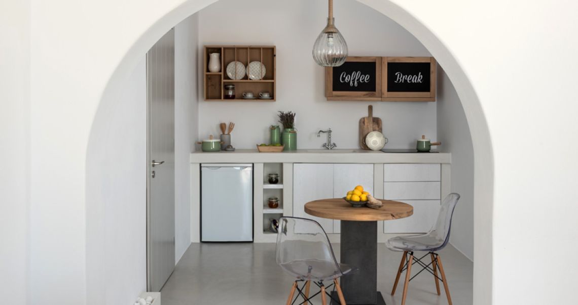 View through a white archway into a modern kitchenette and dining area featuring wooden cabinets, a round table, and clear acrylic chairs.
