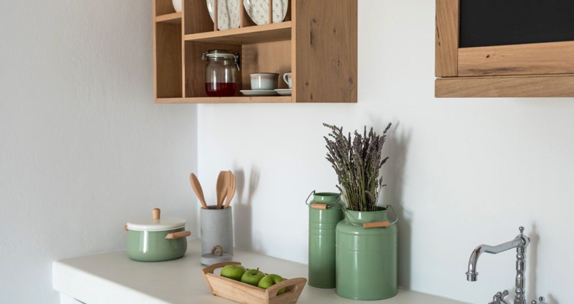 Detail of a kitchen counter styled with sage green cookware, wooden utensils, and open wooden shelving displaying ceramic dishware.